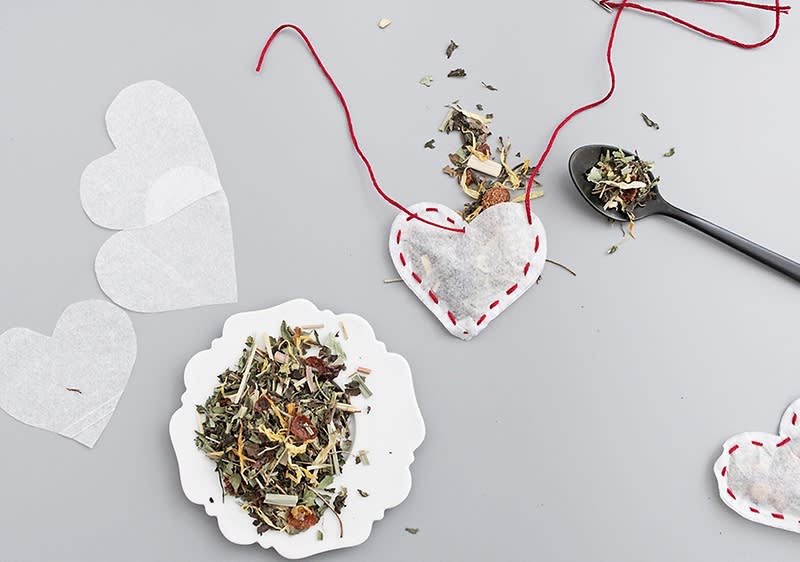 Herzförmige Teebeutel aus Papier, gefĂŒllt mit KrĂ€utern, liegen auf einem grauen Hintergrund. Ein schwarzer Löffel mit KrĂ€utern daneben.