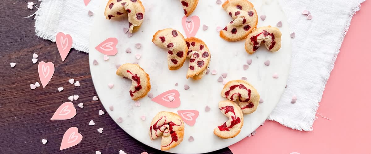 Mehrere Kekse mit roten Flecken liegen auf einem runden weißen Teller, umgeben von kleinen rosa und weißen Herzdekorationen.