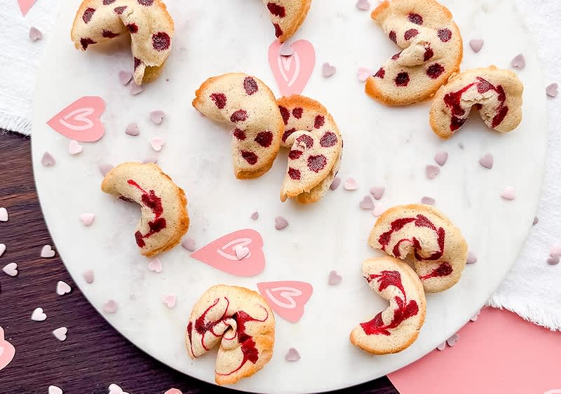 Mehrere halbmondförmige kleine Gebäckstücke mit roten Herz- oder Rosenfüllungen auf weißer Marmorplatte, dekoriert mit kleinen rosa Herzstreuseln und Papierherzen