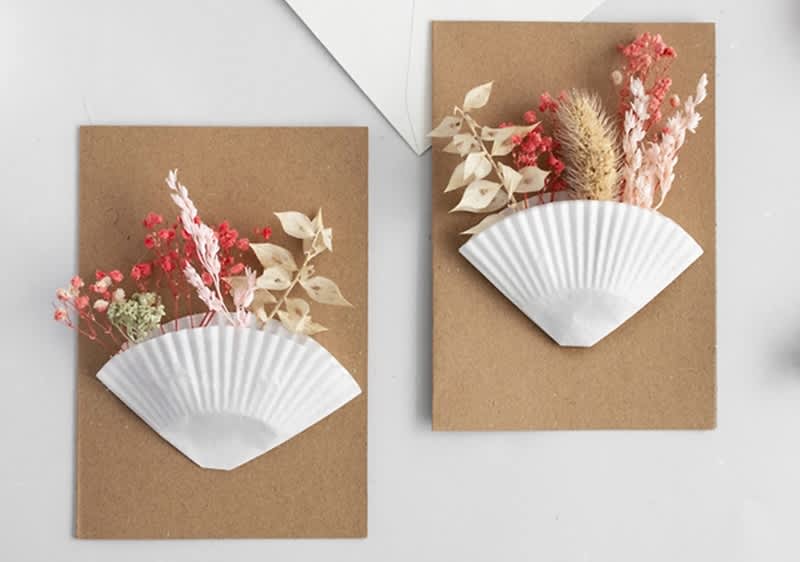 Zwei braune Karten mit weißen Papierfächern, gefüllt mit getrockneten Blumen in Rot, Beige und Weiß.