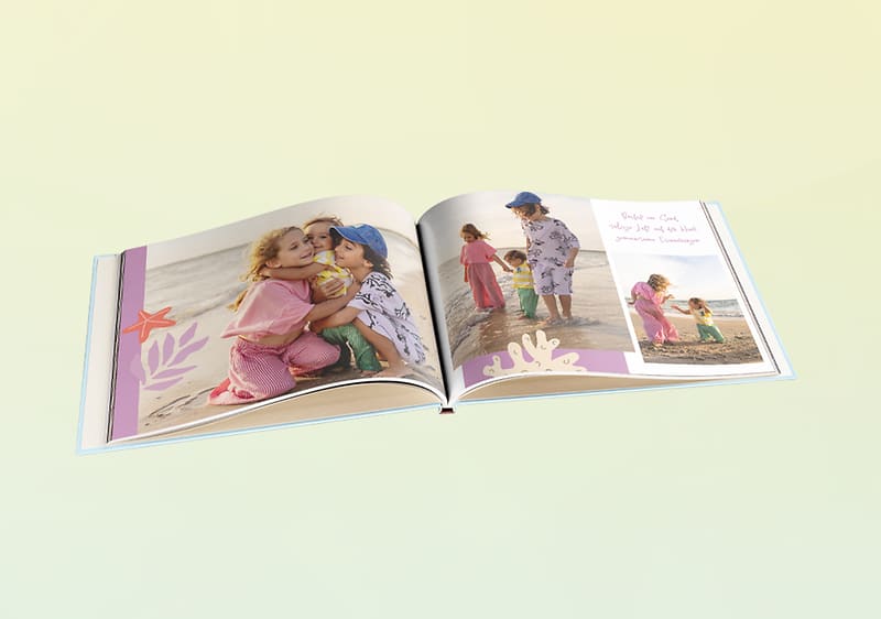 Aufgeschlagenes Fotobuch mit Strandaufnahmen von zwei Kindern, links ein Mädchen, das ein Kleinkind umarmt, rechts mehrere Fotos derselben Kinder am Meer sowie dekorative Muscheln und Textfelder