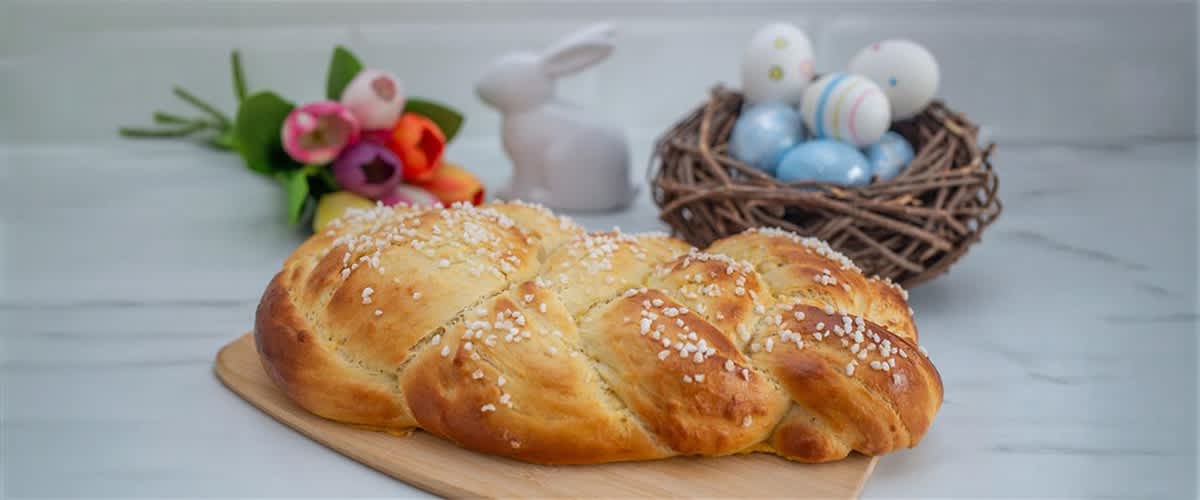 Zopf aus glänzend gebackenem Hefeteig mit Hagelzucker auf einem Holzbrett, im Hintergrund Osterdeko mit gefärbten Eiern im Nest, Keramikhase und Tulpenstrauß