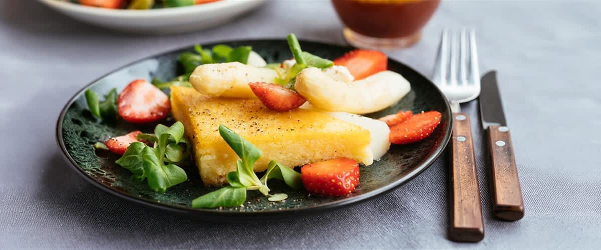 Ein schwarzer Teller mit gebratener Polenta, karamellisierten Bananenscheiben, halbierten Erdbeeren und grünen Sprossen garniert, Besteck rechts daneben