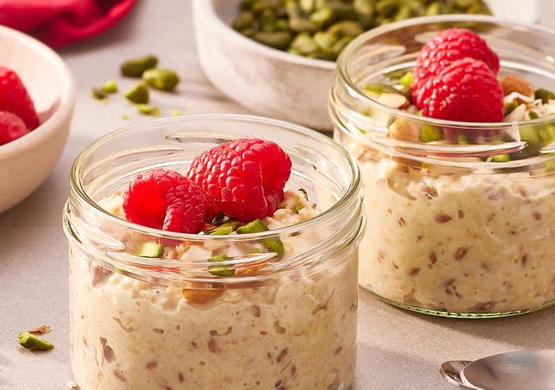 Zwei kleine Glasgläser mit Haferpudding, garniert mit gehackten Pistazien und frischen Himbeeren, nebeneinander auf hellem Untergrund