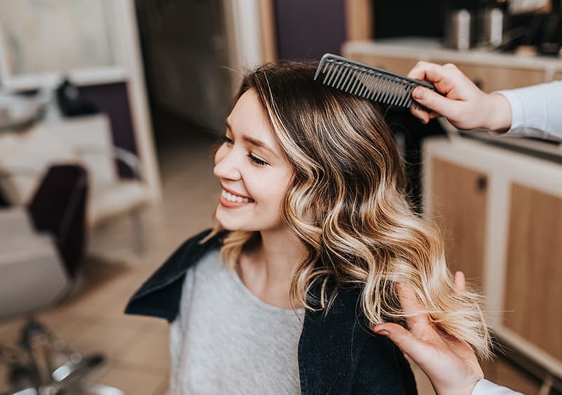 Hand einer Friseurin kämmt welliges, blond gesträhntes Haar einer Kundin in einem Salon