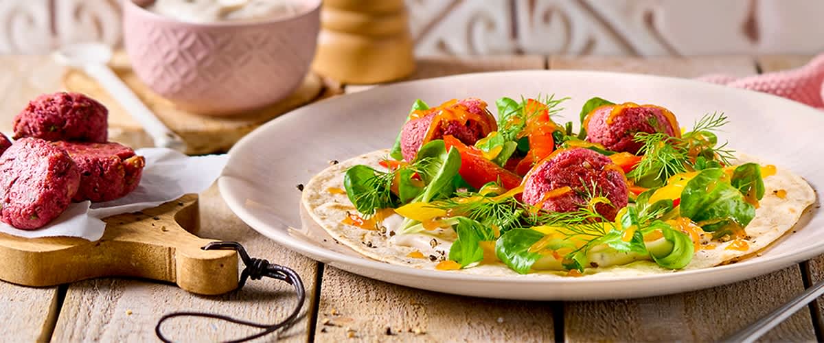 Weiche Tortilla mit Joghurtcreme, darauf Rote-Bete-Bällchen, gedünstete Paprikastreifen, Feldsalat und Dill garniert, serviert auf hellem Teller auf Holzuntergrund
