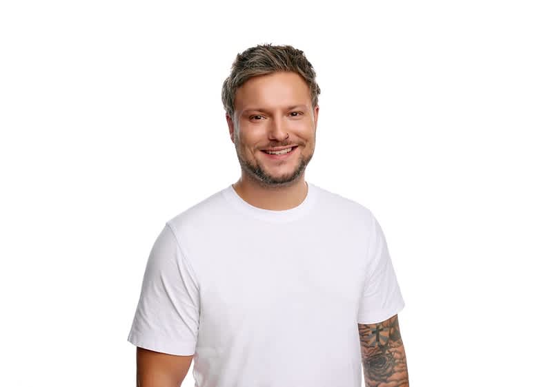 Mann steht vor weißem Hintergrund, trägt weißes T-Shirt und zeigt tätowierten rechten Unterarm, Gesicht unkenntlich gemacht