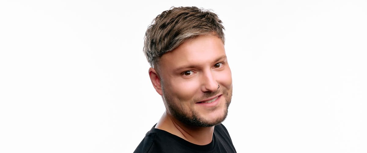 Junger Mann in schwarzem T-Shirt steht vor weißem Hintergrund, Oberkörper leicht zur Seite gedreht, Gesicht unkenntlich gemacht