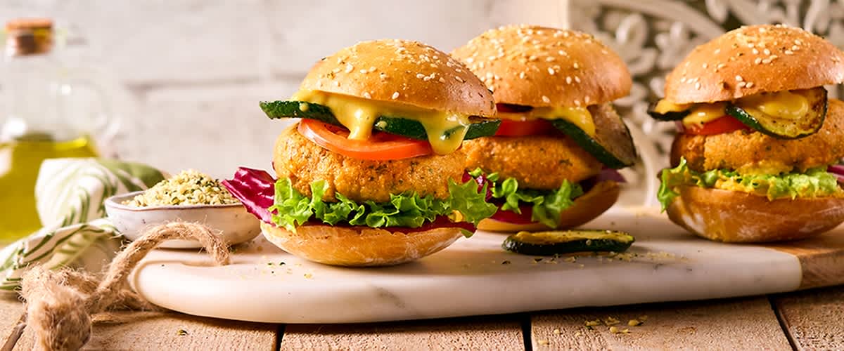 Zwei vegetarische Burger mit Sesambrötchen, knuspriger Panade, Tomatenscheiben, Salat, gegrillter Zucchini und schmelzendem Käse auf einem weißen Servierbrett