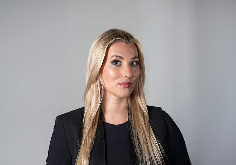 Vorderansicht einer stehenden Person mit langen, blonden Haaren, schwarzem Oberteil und schwarzem Blazer vor neutralem hellgrauem Hintergrund