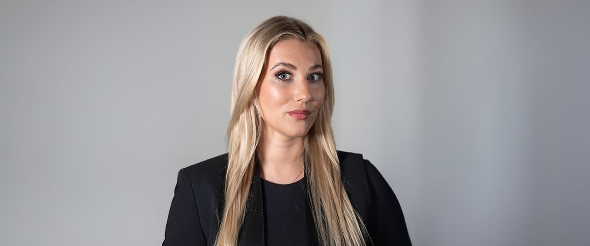 Vorderansicht einer stehenden Person mit langen, blonden Haaren, schwarzem Oberteil und schwarzem Blazer vor neutralem hellgrauem Hintergrund