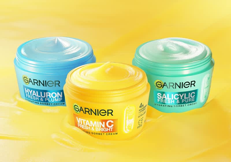 Drei geöffnete Garnier-Gesichtspflege-Tiegel auf gelbem Hintergrund: links blaues Tiegel mit Aufschrift „Hyaluron Fresh & Plump“, vorne gelber Tiegel mit Aufschrift „Vitamin C Fresh & Bright“, rechts grünes Tiegel mit Aufschrift „Salicylic Fresh & Pure“, alle mit sichtbar cremiger Konsistenz