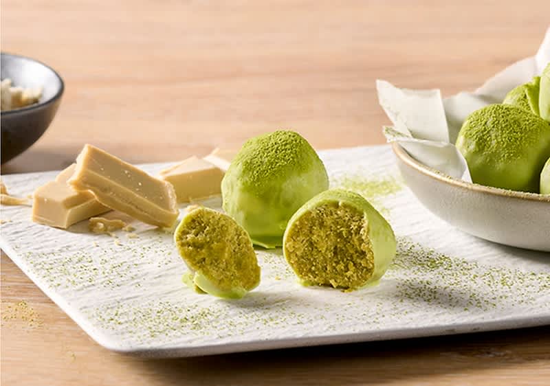 Drei grüne Matcha-Pralinen auf weißem Teller, eine halbiert und innen krümelig, daneben weiße Schokolade in Stücken und Matchapulver bestreut