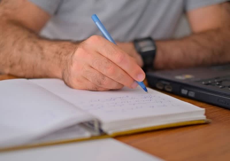 Männliche Hand mit blauem Stift schreibt Notizen in ein aufgeschlagenes Notizbuch neben einem Laptop