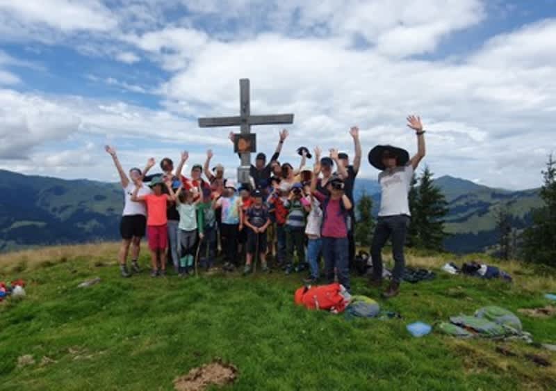 Gruppe von Kindern und zwei Erwachsenen steht auf einem Gipfel vor einem Holzkreuz, viele winken in die Kamera, Berglandschaft im Hintergrund