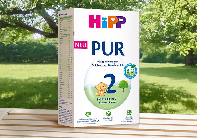 HiPP PUR Folge 2 Bio-Milchpulver-Packung in Weiß mit buntem HiPP-Logo, großer blauer Zahl 2, Aufschrift „PUR“ und „NEU“ sowie Bio-Siegel auf grünem Gartentisch ohne Punkt