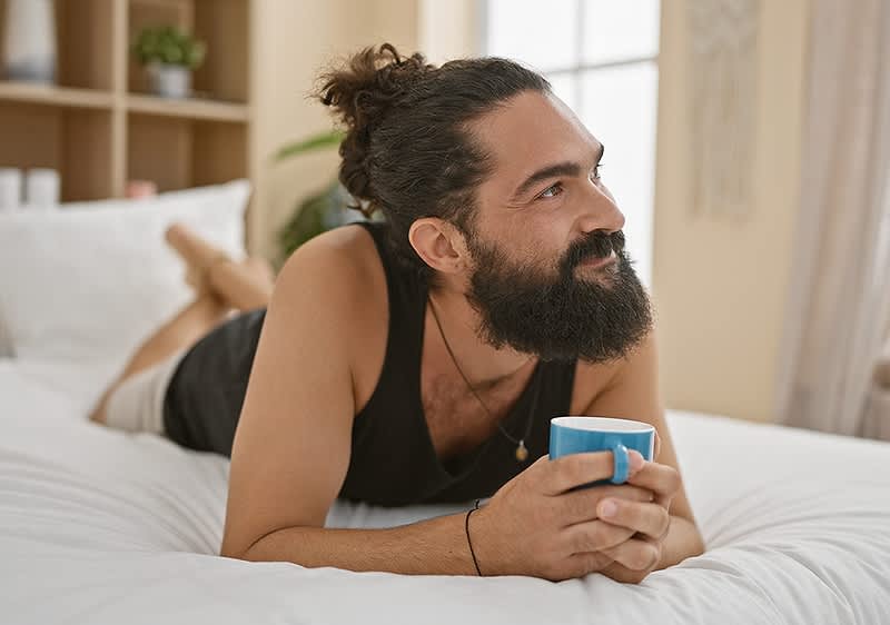 Person liegt auf dem Bauch auf einem Bett, hält eine blaue Tasse mit beiden Händen und blickt zur Seite
