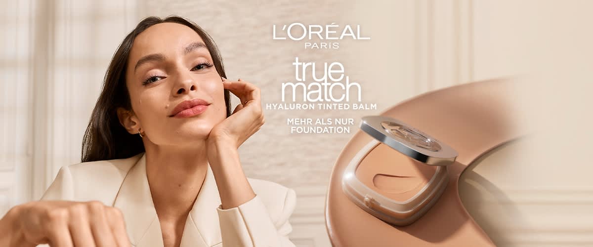 Werkt rotierende Frau in hellem Blazer hält Hand an Kinn, rechts große L'Oréal Paris true match Hyaluron Tinted Balm Kompaktverpackung mit geöffnetem Spiegel und Produkttext Mehr als nur Foundation
