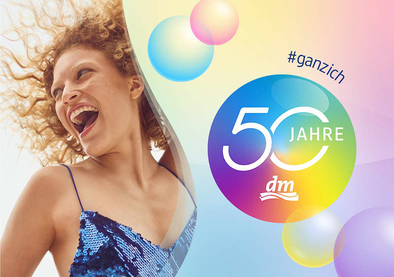 Frau in blau schimmerndem Paillettenkleid mit lockigem Haar, daneben bunter Kreis mit Text „50 Jahre“ und dm-Logo, Hashtag „#ganzich“