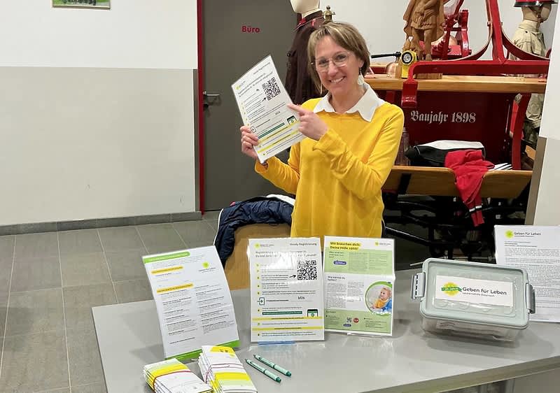 Person in gelbem Pullover steht hinter einem Infotisch und hält ein Flugblatt mit QR-Code, auf dem Tisch Broschüren, Informationsblätter, zwei grüne Kugelschreiber und eine beschriftete Box mit Aufkleber für Spenden oder Rückgabematerialien