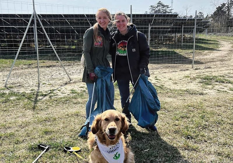 Zwei Personen in Freizeitkleidung stehen auf einer Wiese und halten je einen blauen Müllsack, davor sitzt ein Hund mit einem weißen Halstuch mit grünem Motiv, im Hintergrund Zaun und Solarpaneele