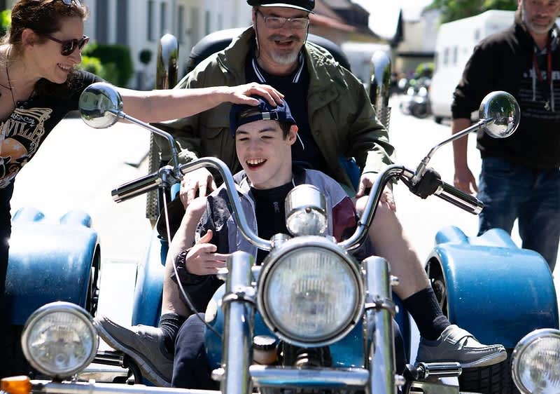 Kind lächelnd auf einem blauen Dreirad-Motorrad sitzend, hinter ihm ein Erwachsener mit Hand unterstützend am Lenker, weitere Personen im Hintergrund, sonniger Außenbereich