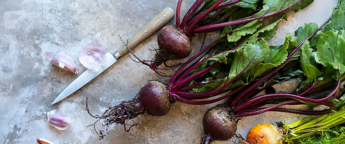 Drei frische Rote Rüben mit Laub liegen auf Stein, links ein Küchenmesser mit Holzgriff, unten rechts eine gelbe Rübe sichtbar