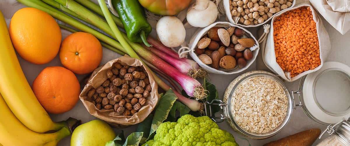 Verschiedene Lebensmittel arrangiert: grüner Blumenkohl, Haferflocken im Glas, Nüsse in Beuteln, Kichererbsen, Pilze, Frühlingszwiebeln, Paprika, Äpfel und Mandarinen