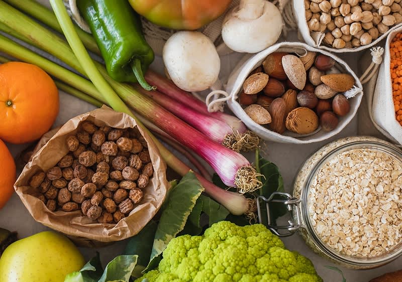 Verschiedene gesunde Lebensmittel auf einem Tisch: Mandarinen, grüner Apfel, Frühlingszwiebeln, Pilze, Tomate, Blumenkohl, Haferflocken im Glas, Nüsse in Stoffbeutel und Kichererbsen in Papierbeutel