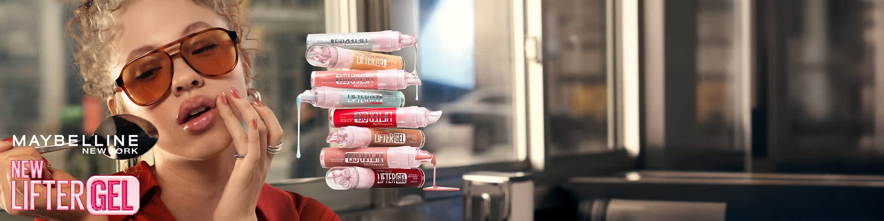A Stapel verschiedener Maybelline Lifter Gel Lippenprodukte in Pastell- und Rottönen, aus den Öffnungen tropft Gel, links Schriftzug „MAYBELLINE NEW YORK“ und „NEW LIFTER GEL“