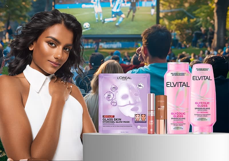 Frau in weißem ärmellosem Oberteil hält Hand an die Schulter, im Vordergrund drei L’Oréal-Produkte: L’Oréal Paris Glas Skin Hydrogel-Maske, Elvital Glycolic Gloss Shampoo und Spülung sowie zwei L’Oréal-Make-up-Stifte, im Hintergrund Menschen sehen ein Fußballspiel auf einer Großbildleinwand