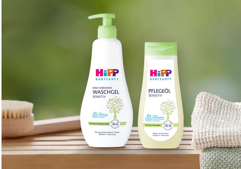 Zwei HiPP Babysanft Produkte auf Holzablage: links weiße Pumpflasche „Haut und Haar Waschgel Sensitiv“, rechts gelbliche Flasche „Pflegeöl Sensitiv“, beide mit grünem Blatt-Motiv und grünen Verschlüssen, rechts daneben gestapelte Handtücher