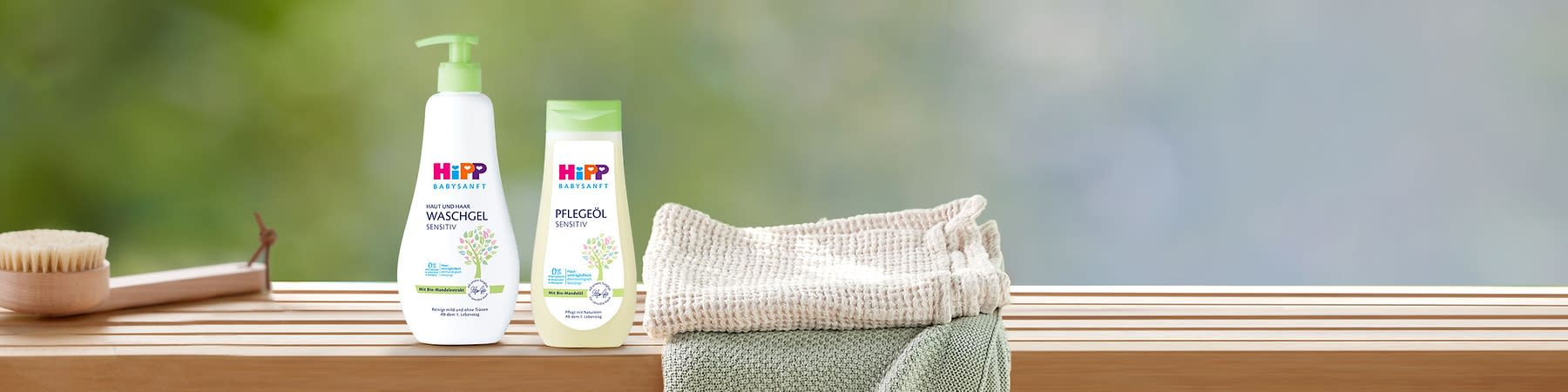 Zwei HiPP Babysanft Produkte auf Holzablage: links weiße Pumpflasche „Haut und Haar Waschgel Sensitiv“, rechts gelbliche Flasche „Pflegeöl Sensitiv“, beide mit grünem Blatt-Motiv und grünen Verschlüssen, rechts daneben gestapelte Handtücher