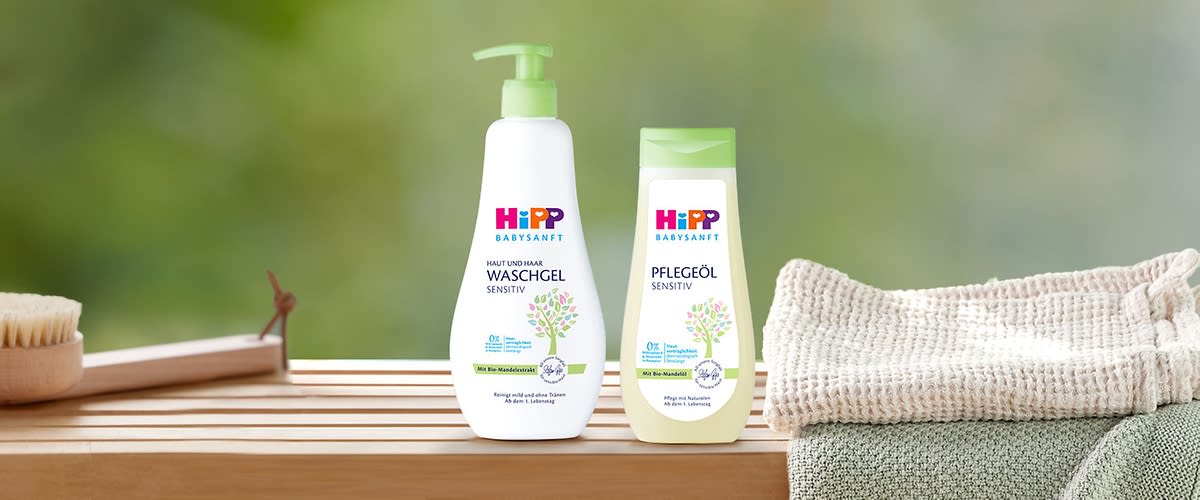 Zwei HiPP Babysanft Produkte auf Holzablage: links weiße Pumpflasche „Haut und Haar Waschgel Sensitiv“, rechts gelbliche Flasche „Pflegeöl Sensitiv“, beide mit grünem Blatt-Motiv und grünen Verschlüssen, rechts daneben gestapelte Handtücher