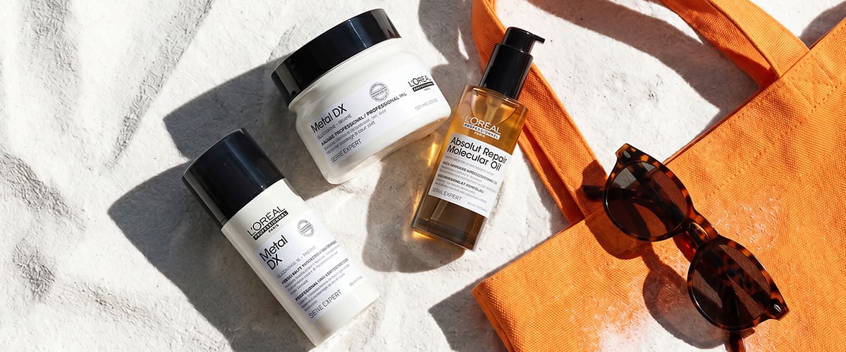 Drei L’Oréal Professional Haarpflegeprodukte auf hellem Steinuntergrund: links eine weiße Flasche „Metal DX“, oben ein weißes Tiegelglas „Metal DX“, rechts ein Pumpspender „Absolut Repair Molecular Oil“, daneben eine orange Stofftasche und eine Sonnenbrille