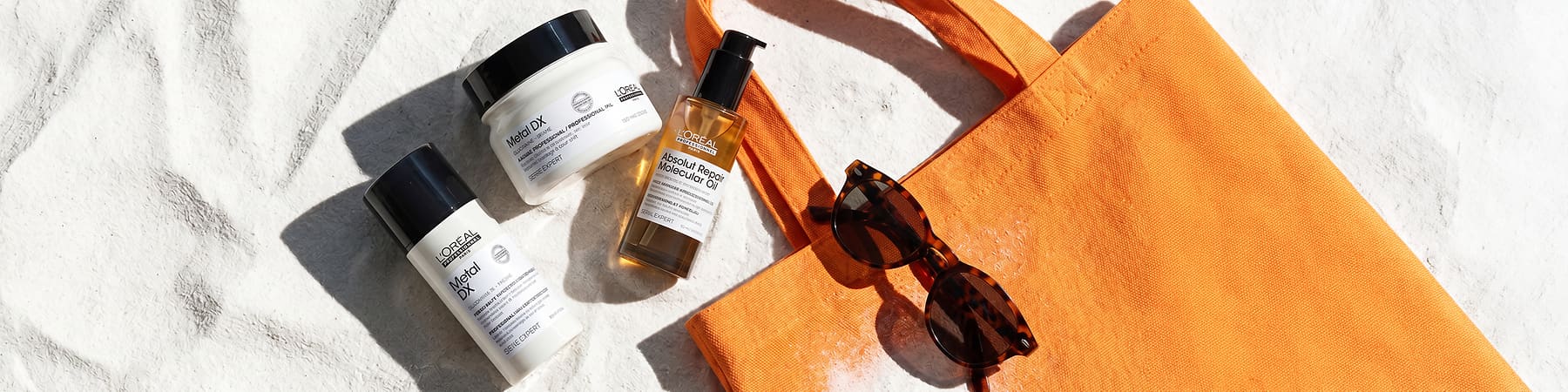 Drei L’Oréal Professional Haarpflegeprodukte auf hellem Steinuntergrund: links eine weiße Flasche „Metal DX“, oben ein weißes Tiegelglas „Metal DX“, rechts ein Pumpspender „Absolut Repair Molecular Oil“, daneben eine orange Stofftasche und eine Sonnenbrille
