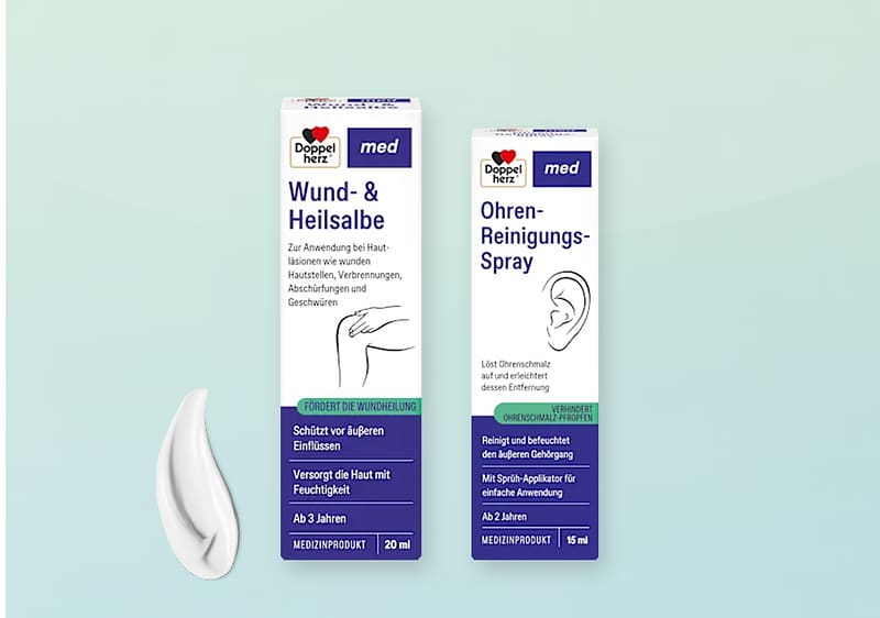 Zwei weiße Medikamentenverpackungen der Marke Doppelherz med vor hellem Verlaufshintergrund: links „Wund- & Heilsalbe“ 20 ml mit grünem Streifen und Piktogramm einer Hand, rechts „Ohren-Reinigungs-Spray“ 15 ml mit blauem Streifen und Ohr-Illustration, darunter links eine aufgetragene Salbenportion