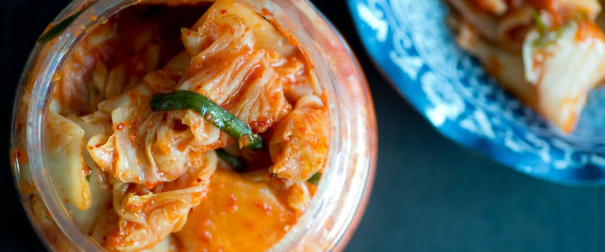Ein offenes Glas Kimchi mit roten Gewürzflocken, daneben ein blau-weißes Schälchen mit Kimchi und rechts liegende Essstäbchen auf dunklem Untergrund