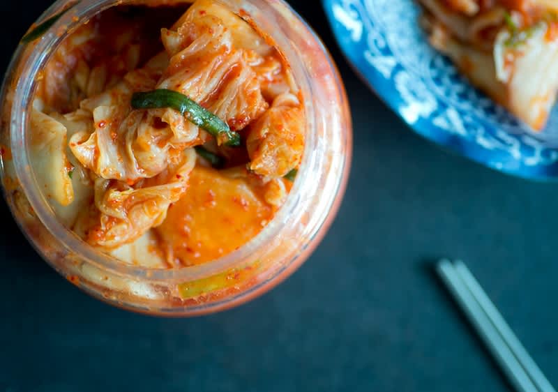 Offenes Glas Kimchi mit sichtbaren Chinakohlstücken in roter Gewürzpaste, rechts ein blau-weiß gemustertes Schälchen mit Kimchi und unten rechts Essstäbchen auf dunklem Untergrund