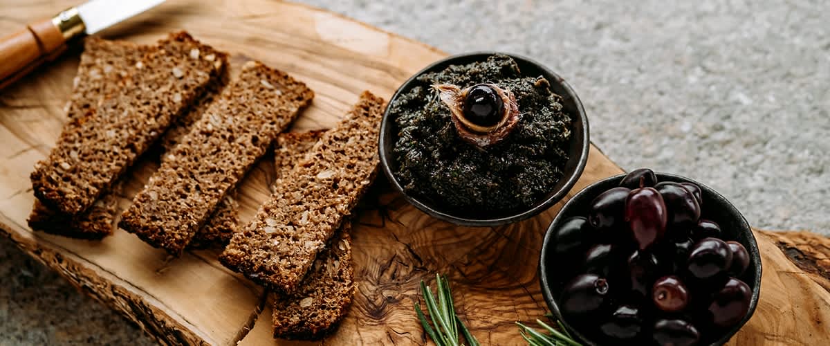 Aufgeschnittener dunkler Vollkornbrot auf einem rustikalen Olivenholzbrett, daneben eine Schale schwarzer Oliven, eine Schale Olivenpaste mit einer Olive obenauf und Rosmarinzweige, Messer am Brett
