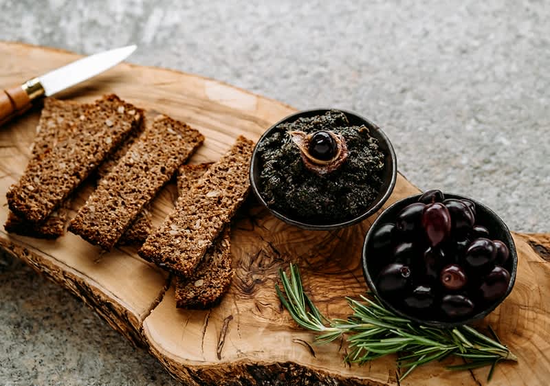 Auf einem rustikalen Olivenholzbrett liegen vier Scheiben dunkles Vollkornbrot, eine Schale mit schwarzen Oliven, eine Schale mit schwarzem Tapenade-Aufstrich garniert mit einer Olive und ein Zweig Rosmarin