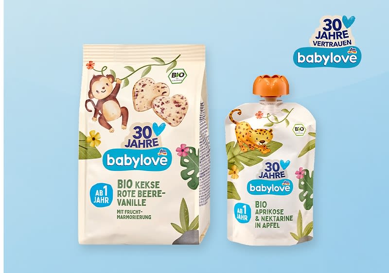 Links eine beige Tüte Babylove Bio-Kekse „Rote Beere-Vanille“ mit Affen-Illustration und Herzkeksen, rechts eine Quetschpackung Babylove Bio „Aprikose & Nektarine in Apfel“ mit Leoparden-Illustration, oben rechts kleines Babylove-Logo mit Schriftzug „30 Jahre Vertrauen“