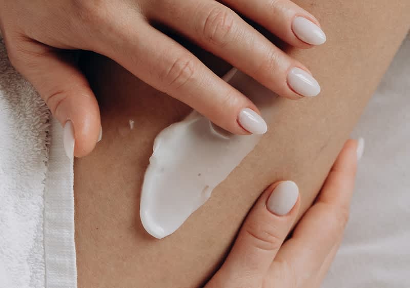 Hand mit gepflegten, hell lackierten Nägeln trägt eine weiße Creme auf Hautstelle auf, darunter ein weißes Handtuch sichtbar