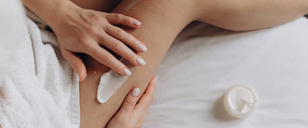 Hände mit gepflegten, hell lackierten Nägeln verteilen eine Creme auf dem Oberschenkel, weißer Frottéhandtuchsaum sichtbar