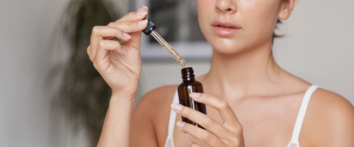 Eine Frau hält eine braune Glasflasche mit Pipette und lässt ein paar Tropfen eines öligen Serums in die Flasche fallen, sichtbarer Oberkörper in weißem Trägertop ohne Hintergrunddetails