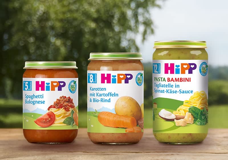 Drei Hipp-Babynahrungs-Gläser auf Holztisch: links Glas mit roter Spaghetti-Bolognese und Aufschrift „5+ Spaghetti Bolognese“, mittig Glas mit Karotten, Kartoffel und Bio-Rind und Aufschrift „8+ Karotten mit Kartoffeln & Bio-Rind“, rechts Glas mit grüner Spinat-Käse-Sauce und Aufschrift „12+ Pasta Bambini Tagliatelle in Spinat-Käse-Sauce“