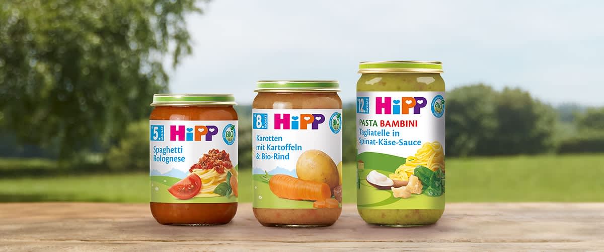 Drei Hipp-Babynahrungs-Gläser auf Holztisch: links Glas mit roter Spaghetti-Bolognese und Aufschrift „5+ Spaghetti Bolognese“, mittig Glas mit Karotten, Kartoffel und Bio-Rind und Aufschrift „8+ Karotten mit Kartoffeln & Bio-Rind“, rechts Glas mit grüner Spinat-Käse-Sauce und Aufschrift „12+ Pasta Bambini Tagliatelle in Spinat-Käse-Sauce“