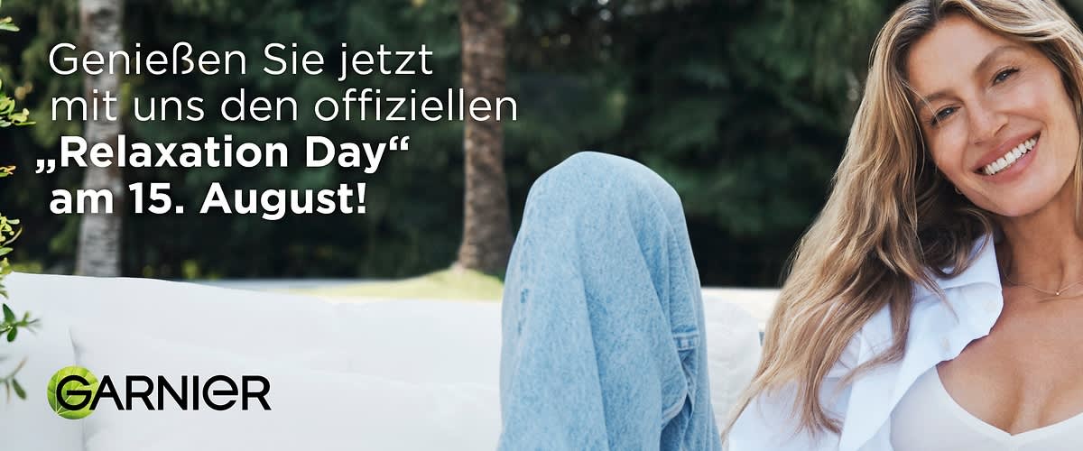 Lächelnde Frau mittleren Alters sitzt entspannt, trägt weiße Bluse offen über weißem Top und feines Halskettchen, lange wellige Haare fallen über die Schultern, verschwommener grüner Hintergrund