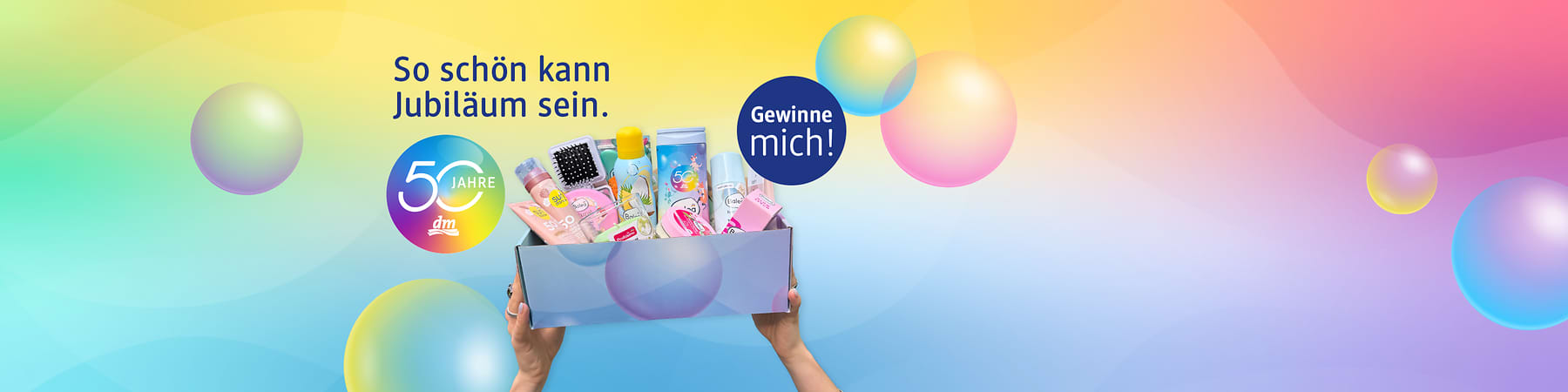 Bunte Geschenkbox mit Pflegeprodukten und Überraschungen, gehalten von zwei Händen, links Slogan „So schön kann Jubiläum sein.“, darunter rundes Logo „50 Jahre dm“, rechts blauer Kreis mit Text „Gewinne mich!“