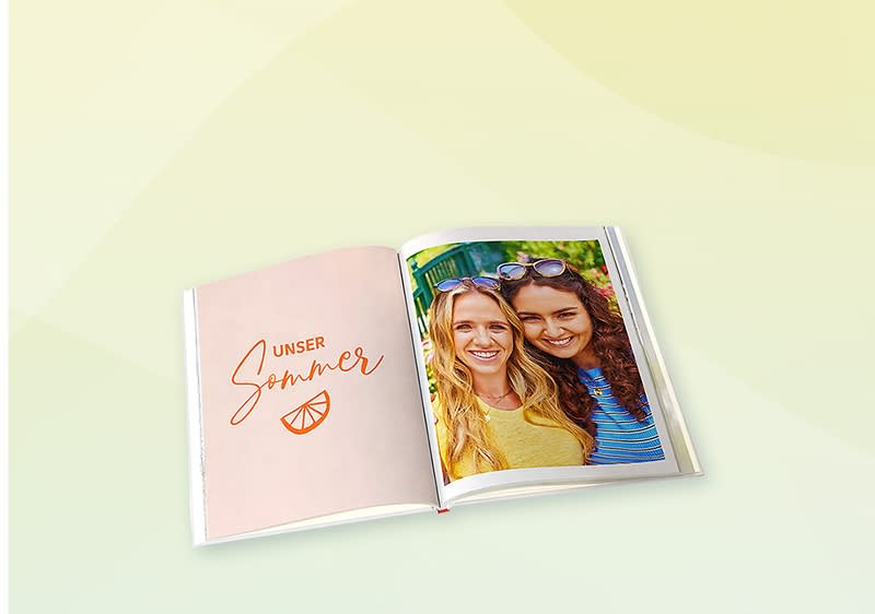 Aufgeschlagenes Fotobuch: linke Seite rosa mit Schrift „Unser Sommer“ und einer Orangen-Scheibe, rechte Seite Foto zweier lächelnder Frauen mit Sonnenbrille auf dem Kopf, helle Hintergrundfarbe